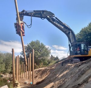 A gray John Deere excavator equipped with a Gilbert MG-90 vibratory hammer drives a wooden pile into the ground along a riverbank to create a retaining structure.
